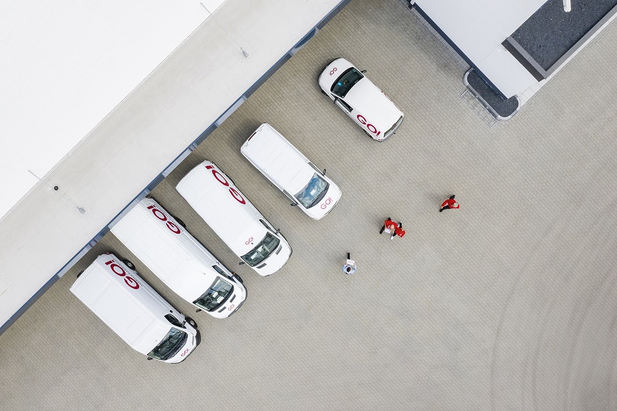 Aus Vogelperspektive ist ein Ausschnitt des Hubs bei Tag zu sehen. Vor dem Hub parken GO! Transporter mit dem Lagerraum zu den Einganstoren des Hubs gedreht, bereit für die Beladung. Vor den GO! Transportern gehen GO! Kuriere auf Ihre Transporter zu