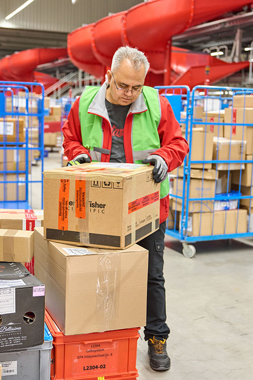 Ein Mitarbeiter (m/w/d) von GO! Express & Logistics steht im Lager. Er trägt Sicherheitsschuhe, eine rote Jacke und darüber eine Warnweste. Vor ihm liegen ein paar Pakete. Er liest den Versandaufkleber um die Sendung entsprechend zum weiteren Transport zu sortieren. Im Hintergrund sieht man weitere Rollwägen mit anderen Sendungen.