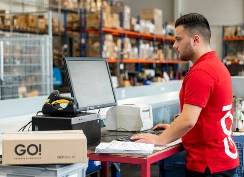 Mitarbeiter arbeiten im Lager. Einer kontrolliert die Sendungen mithilfe eines Computers, während andere im Hintergrund Pakete in ein Lieferfahrzeug laden.