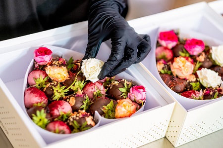 Zwei herzgeformte Schachteln voll mit verschieden überzogenen und verzierten Erdbeeren und Rosen liegen nebeneinander. Eine Hand mit schwarzem hygiene Handschuh legt eine weitere weiße Rose in die vordere Schachtel