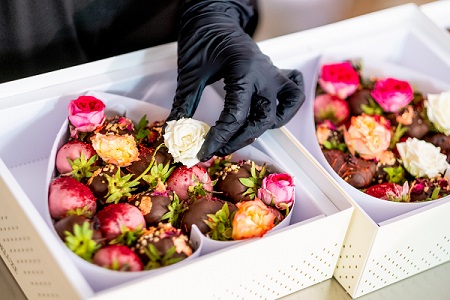 Frailice Herzschachteln Zwei herzgeformte Schachteln voll mit verschieden überzogenen und verzierten Erdbeeren und Rosen liegen nebeneinander. Eine Hand mit schwarzem hygiene Handschuh legt eine weitere weiße Rose in die vordere Schachtel