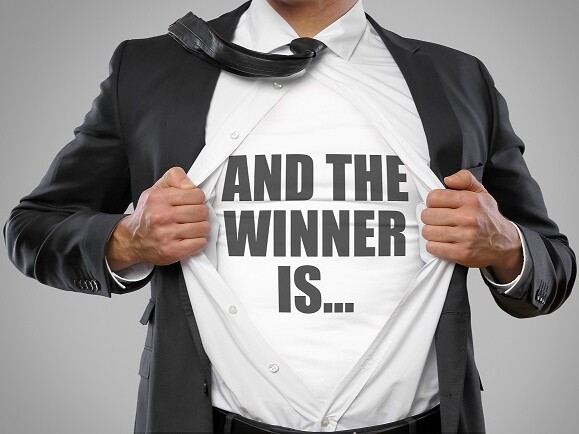 And The Winner Is... Nahaufnahme des Körpers, gesicht nicht zu sehen. Ein Mann im schwarzen Anzug, mit weißem Hemd und Unterhemd, reißt sein Hemd mit beiden Händen auf. Die schwarze Krawatte liegt nach hinten über seine rechte Schulter. Auf dem Unterhemd steht in Großbuchstaben And the winner is...