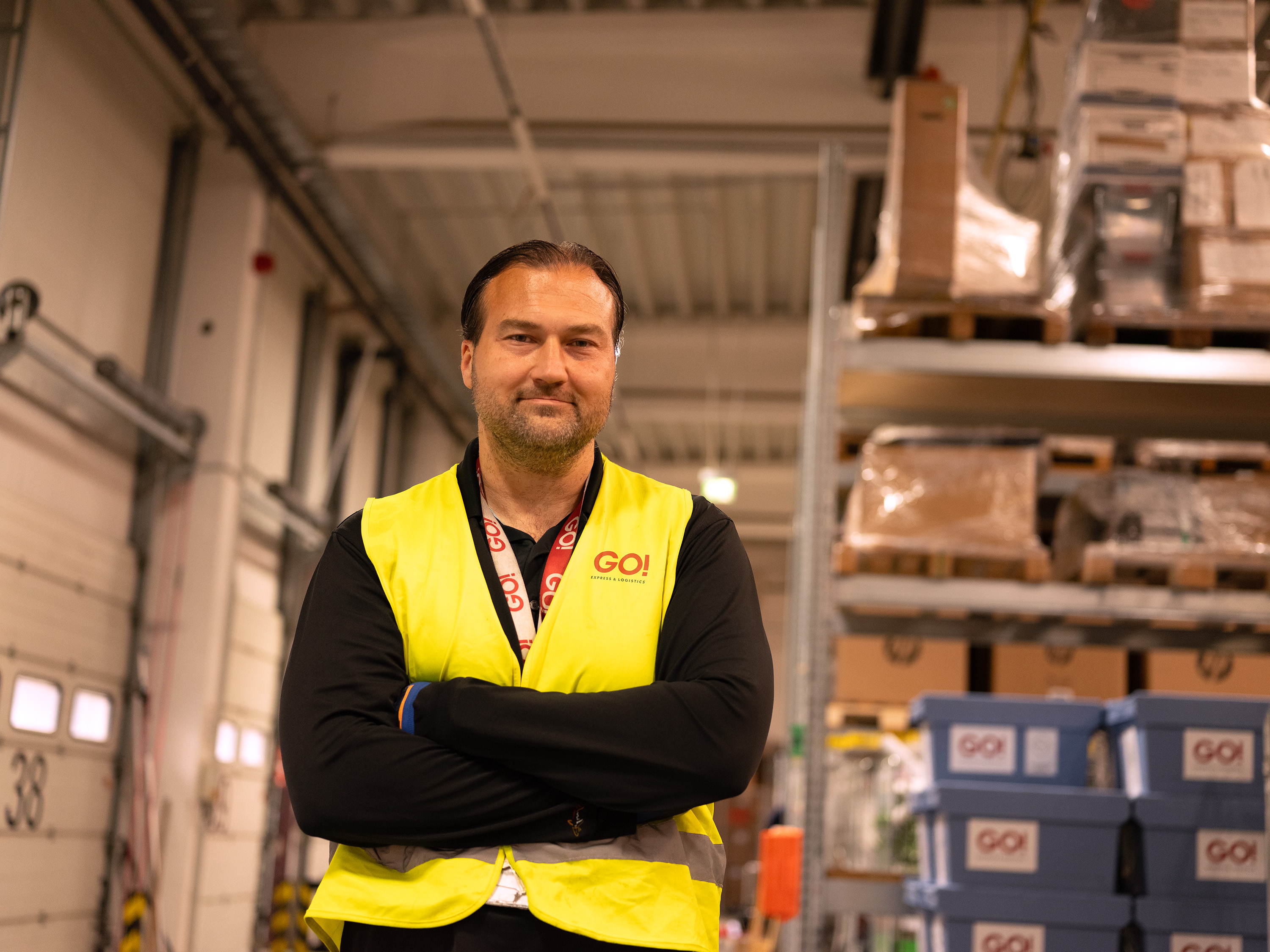Ein_e Mitarbeiter_in von GO! Express & Logistics steht im Lager, trägt eine gelbe Warnweste. Im Hintergrund sieht man mehrere Regale in den Sendungen liegen, die bereits für den weiteren Transport verteilt sind.