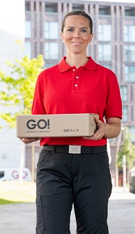 Lächelnde GO! Kurierin mit kleinem GO! Paket in der Hand vor einem Bürogebäude