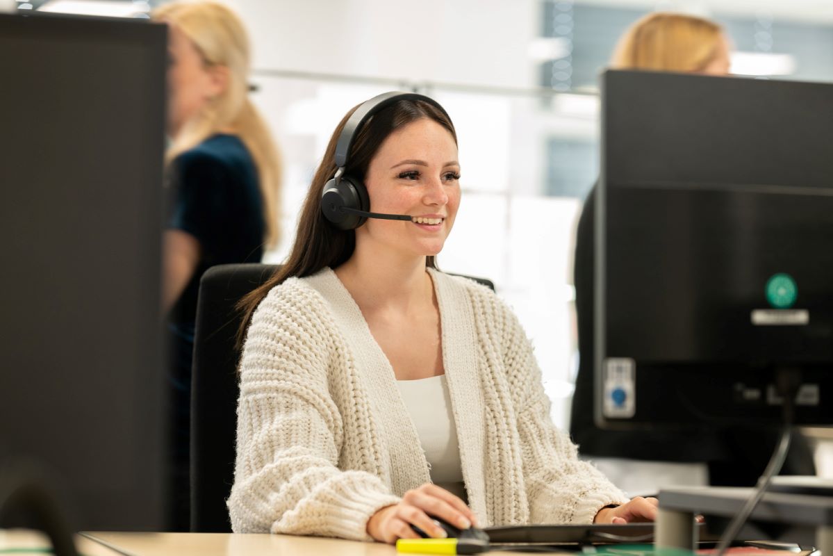 Ein_e kaufmännische_r Mitarbeiter_in von GO! Express & Logistics GmbH sitzt vor dem PC, trägt ein Headset und spricht gerade. Auf dem Schreibtisch sieht man einen Monitor. Die Hände sind auf der Tastatur. Im Hintergrund sieht man zwei weitere Kolleg_innen im Büro.