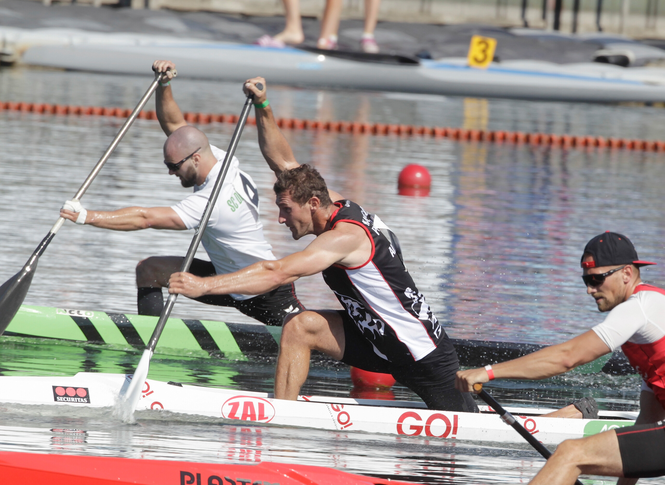 In Seitenaufnahme. Sebastian Brendel Paddel im Wettrennen mit zwei weiteren Kanuten, im Wasser. Auf seinem Kanu das GO! Logo