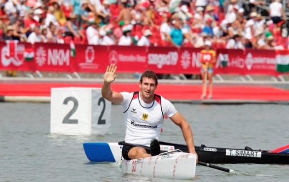 Sebastian Brendel sitz auf seinem Kanu, mit GO! Logo, im Wasser und winkt zur Kamera Sebastian Brendel sitz auf seinem Kanu, mit GO! Logo, im Wasser und winkt zur Kamera. Im Hintergrund ist eine Tribüne voll mit Zuschauern.