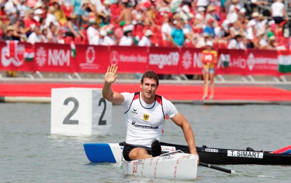 Sebastian Brendel sitz auf seinem Kanu, mit GO! Logo, im Wasser und winkt zur Kamera. Im Hintergrund ist eine Tribüne voll mit Zuschauern.