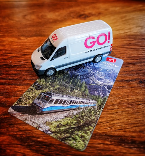 Auf einem Holztisch steht ein miniatur GO! Transporter. Unter dem GO! Transporter ist eine Postkarte von der Zugspitze zu sehen, auf dem ein Zug durch den Wald fährt, im Hintergrund die Zugspitze