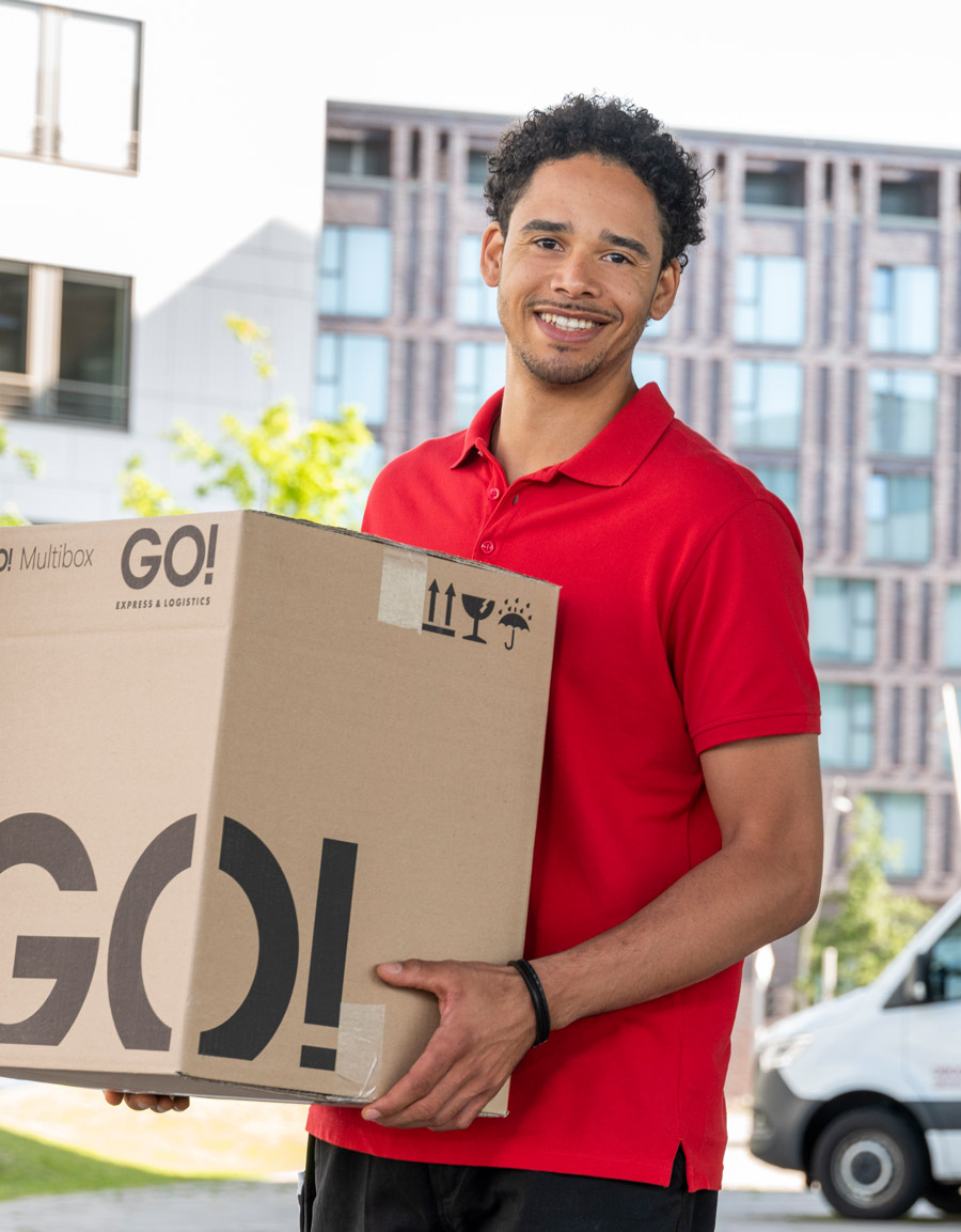 Lächelnder GO! Kurier mit mittelgroßem GO! Paket in der Hand vor einem Bürogebäude