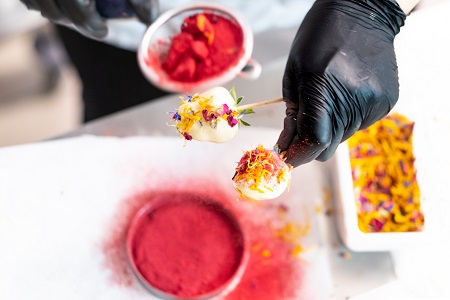 Eine Hand mit schwarzem hygiene Handschuh hält zwei Erdbeeren an Holzstielen. Die Erdbeeren sind mit weißer Schokolade ummantelt und mit gelb und lila Blüten verziert. Im Hintergrund ist ein Sieb mit rotem Pulver verschwommen