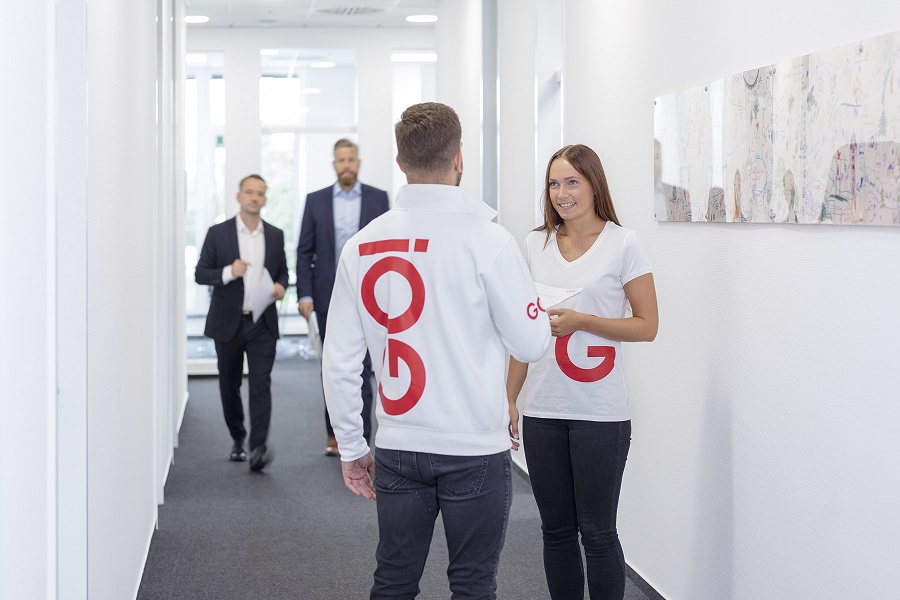 Zwei GO! Mitarbeiter in weißem GO! T-Shirt und GO! Jacke unterhalten sich im Vordergrund, im Flurbereich eines Büros. Im Hintergrund kommen zwei Männer in Anzügen auf sie zu