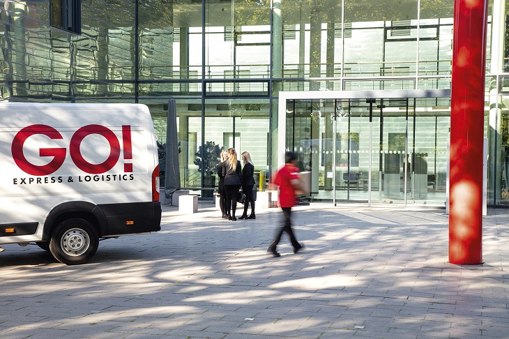 GO! Kurier geht zum Eingang eines Bürogebäudes mit Glaswänden. Links ein GO! Transporter mit dem GO! Logo