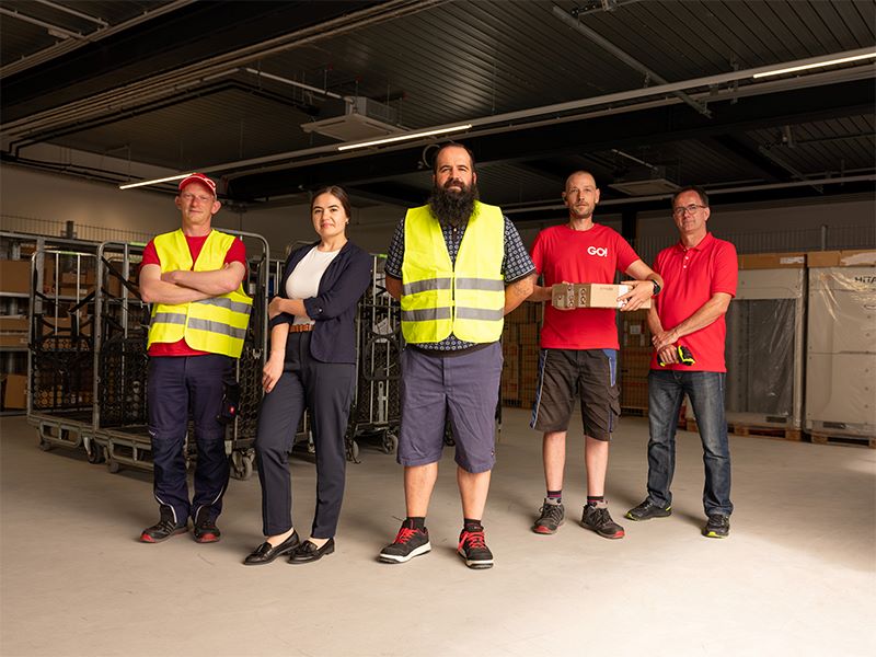 Mitarbeiter (m/w/d) GO! Express & Logistics GmbH 5 Mitarbeiter von GO! Express & Logistics stehen im Lager. Jeder einzelne steht für einen anderen Aufgabenbereich bei GO! Eine davon ist eine Mitarbeiterin. Sie trägt einen Hosenanzug. Zwei andere tragen eine Warnweste und Sicherheitsschuhe. Ein Mitarbeiter in kurzer Hose ist ein Kurierfahrer. Er hält ein Päckchen in der Hand. Der letzte Mitarbeiter hält einen Scanner in der Hand mit dem die Versandaufkleber gescannt werden um dem Kunden immer die Möglichkeit zu geben zu prüfen, wo sich gerade seine Sendung befindet. Alle 5 befinden sich in einer Halle, in der die Sendungen ankommen und für den weiteren Transport verteilt werden. Im Hintergrund sind Rollwägen zu sehen, auf denen die Sendungen zwischengelagert werden.
