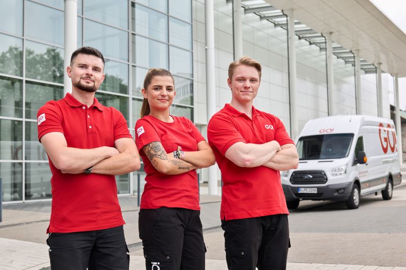 Zwei Fahrer und eine Fahrerin stehen vor einem Gebäude. Hinter den dreien steht ein Transporter von GO!. Das Fahrzeug ist weiß lackiert und mit GO!-Aufklebern in roter Schrift beklebt.
