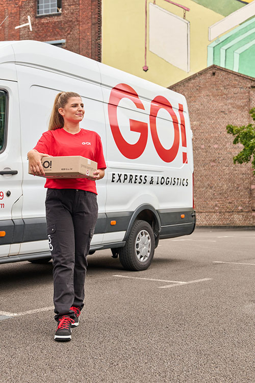 Eine Kurierfahrerin (m/w/d) läuft von ihrem Transporter weg. Sie (m/w/d) trägt eine Sendung in ihrer Hand. Sie trägt Sicherheitsschuhe, eine dunkle Hose und ein rotes GO! T-Shirt