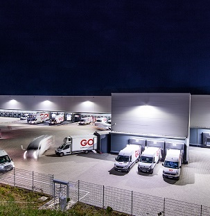 Mehrere GO! Transporter fahren bei Nacht über den Hof des Zentral-Hubs in Niederaula, andere Transporter stehen an den Be- und Entladungstoren.