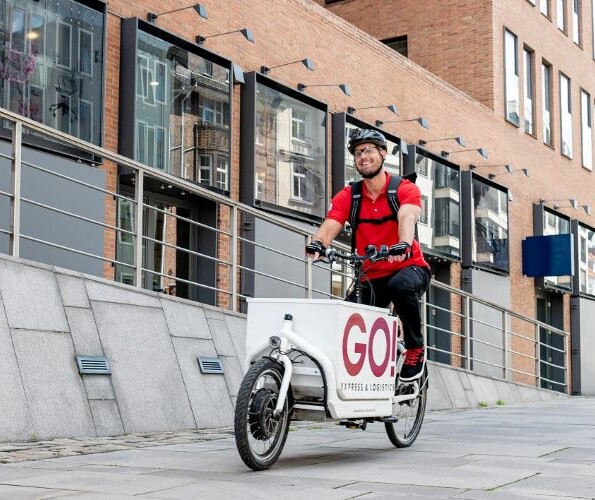 Kurier fährt lächelnd auf einem GO! Lastenfahrrad an einem Haus vorbei.