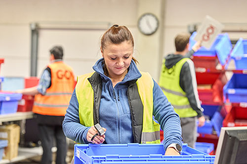 Eine Mitarbeiterin (m/w/d) von GO! Express & Logistics steht im Lager, trägt eine gelbe Warnweste. Im Hintergrund sieht man mehrere Regale in den Sendungen liegen, die bereits für den weiteren Transport verteilt sind.