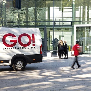 Im Vordergrund steht ein GO! Transporter. Ein GO! Kurier geht mit einem Paket in der Hand vom Auto zu einem Gebäude im Hintergrund. Die Wände des Gebäudes sind aus Glas.