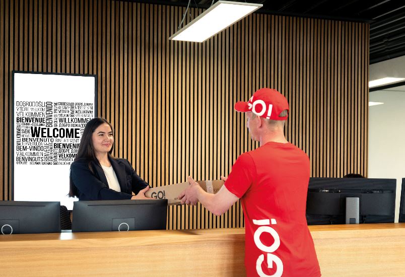 Ein Kurier (m/w/d) von GO! Express & Logistics stellt gerade eine Sendung bei unserem Kunden zu. Er trägt ein rotes T-Shirt mit GO!-Logo