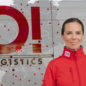 Eine Frau, mit einer roten GO! Jacke steht, zur Kamera lächelnd, vor einer Karte von Deutschland, auf der GO! Stationen mit roten vierecken markiert und durch Striche mit einerander verbunden sind. Über der Karte ist das GO! Logo, leicht transparent, gedruckt.
