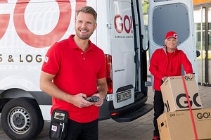 Zwei GO! Kuriere stehen vor einem GO! Transporter. Die Tür zum Lagerraum ist geöffnet. Während ein GO! Kurier zwei Pakete mit einem Sackkarren schiebt, tippt der Andere in sein mobiles computing Gerät. Beide lächeln dabei, der Blick von sich aus nach links gerichtet 