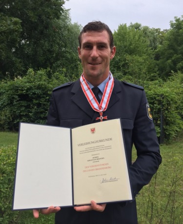 Sebastian Brendel mit Urkunde Sebastian Brendel hält seine Urkunde in die Kamera. Um den Hals trägt er seinen Verdienstorden. Im Hintergrund sind eine Wiese, Bäume und dichte Gebüche zu sehen