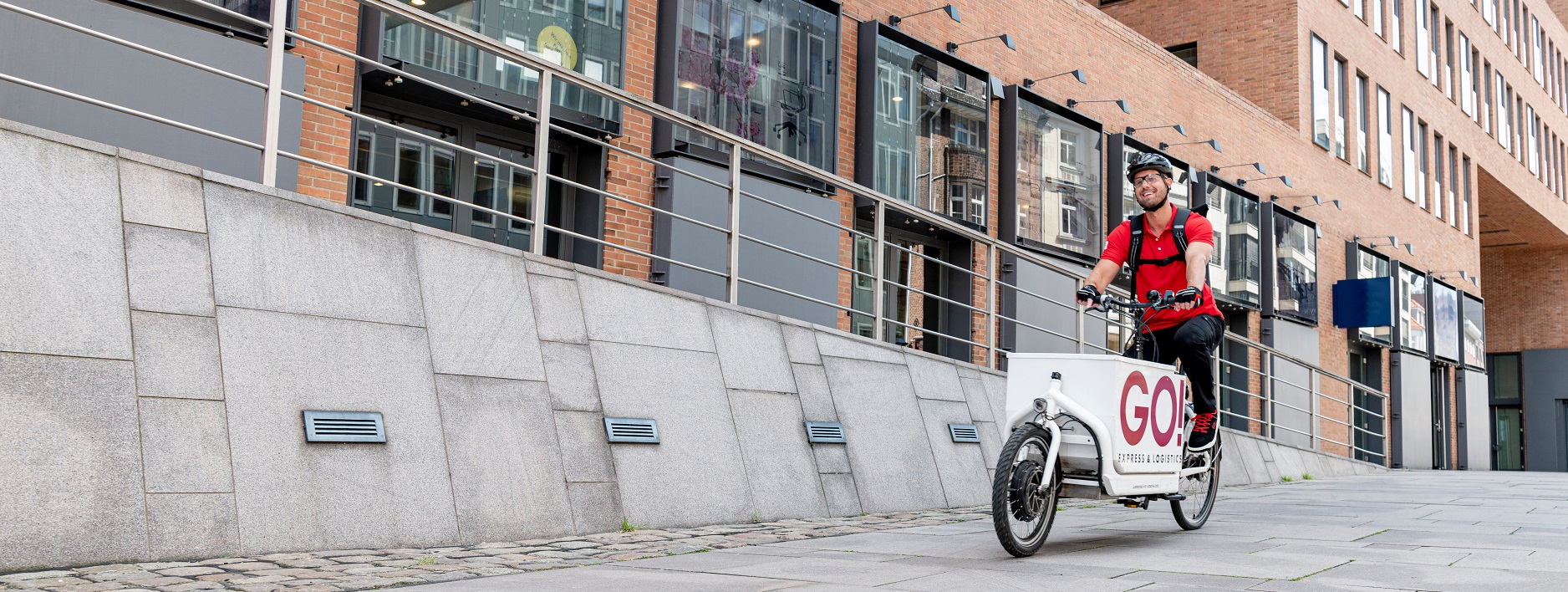 GO! Fahrradkurier fährt auf Lastenrad einen breiten Weg vor einem modernen Backsteingebäude runter