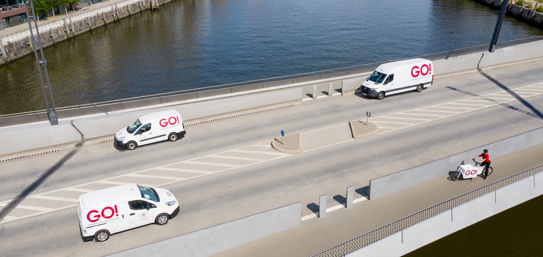 Drei GO! Fahrzeuge und eine Fahrradkurierin auf einem Lastenrad fahren auf beiden Straßenseiten über eine Brücke in Hamburg