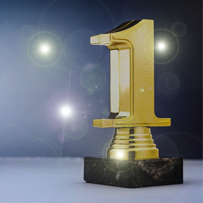 Branchenchampion mit herausragendem Preis- / Leistungsverhältnis Ein goldener Pokal, als die Nummer 1 geformt steht im Vordergrund. Im Hintergrund sind einzelne verschwommene, braune, goldene und weiße Lichter, die wie Konfeti wirken